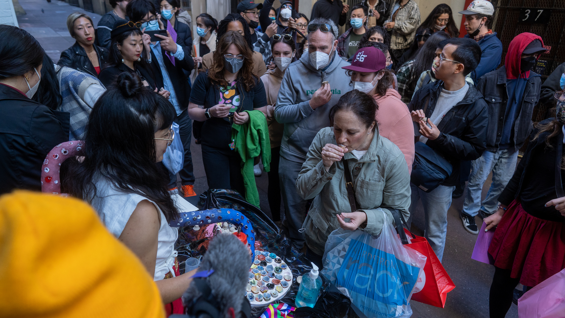 Crowd of people, many masked, waiting in front of table. Artist, wearing a ceramic object, is pouring liquid into a small ceramic cup that a woman is holding.