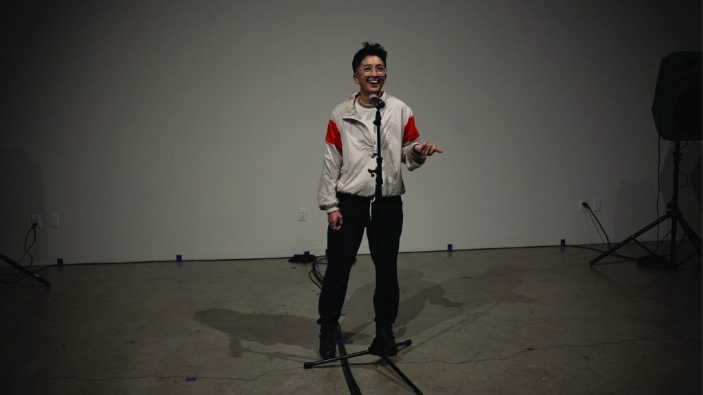 Sun, a person with short black hair sticking up, wearing a sporty lightweight jacket and black paints, standing in front of a mic, smiling and gesturing